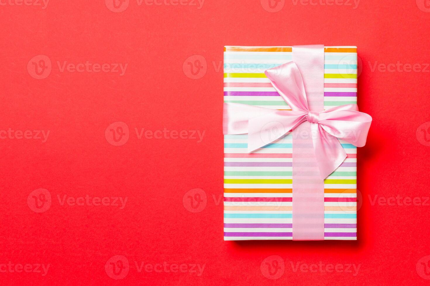 wrapped Christmas or other holiday handmade present in paper with pink ribbon on red background. Present box, decoration of gift on colored table, top view with copy space photo