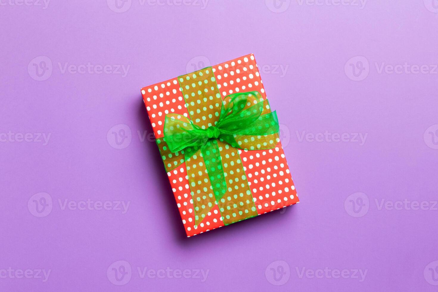 wrapped Christmas or other holiday handmade present in paper with green ribbon on purple background. Present box, decoration of gift on colored table, top view with copy space photo