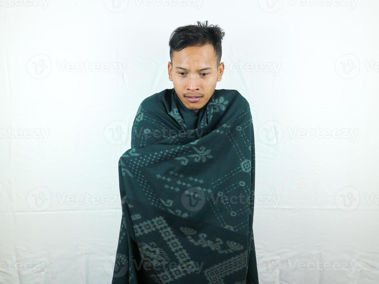 A portrait of happy Asian man feeling cold and covered under a blanket on the bed in the morning. Freshness after waking up from sleep. photo