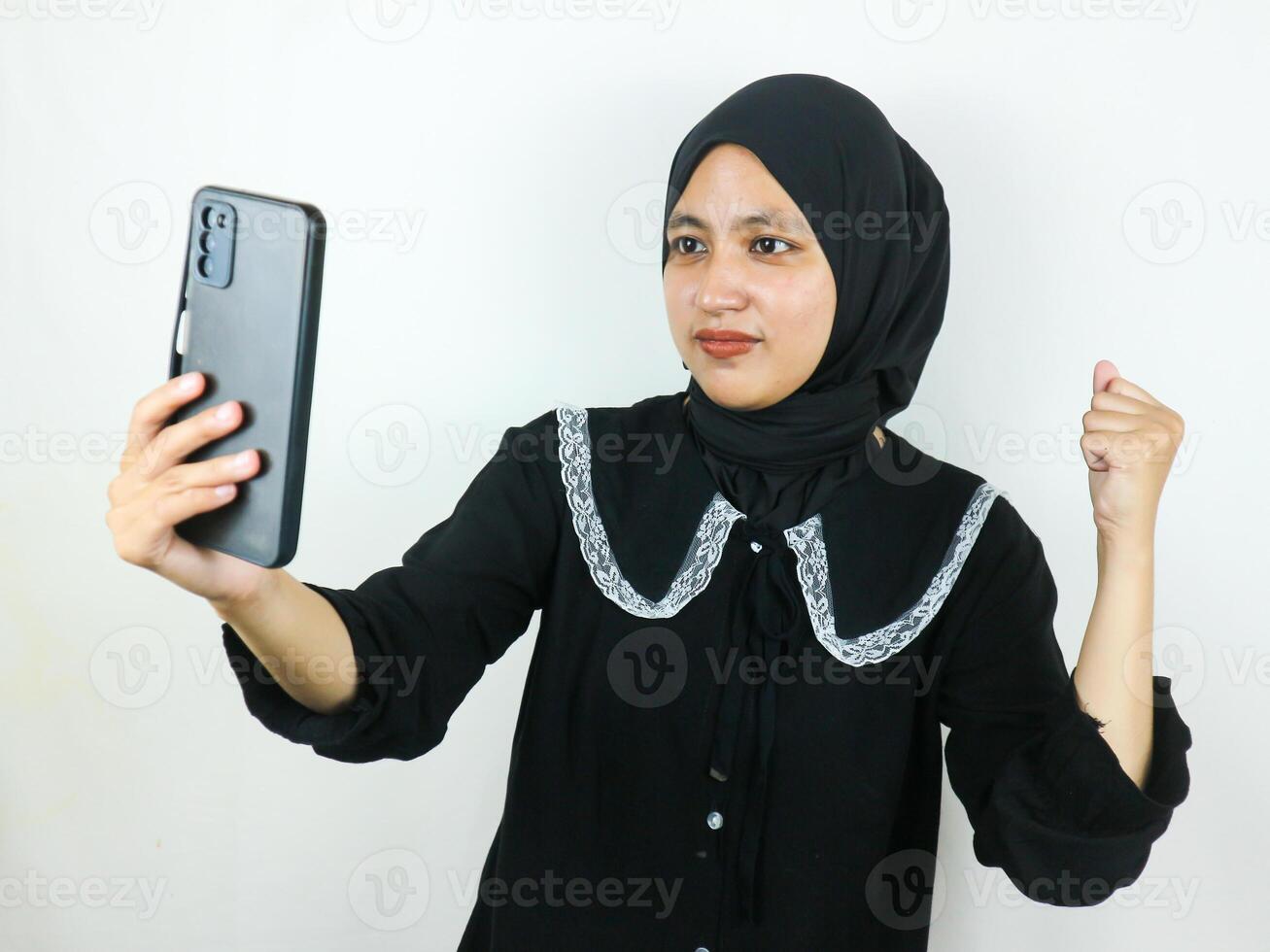 emocionado hermosa asiático mujer en hijab utilizando un móvil teléfono apretando puño recibido bueno Noticias foto