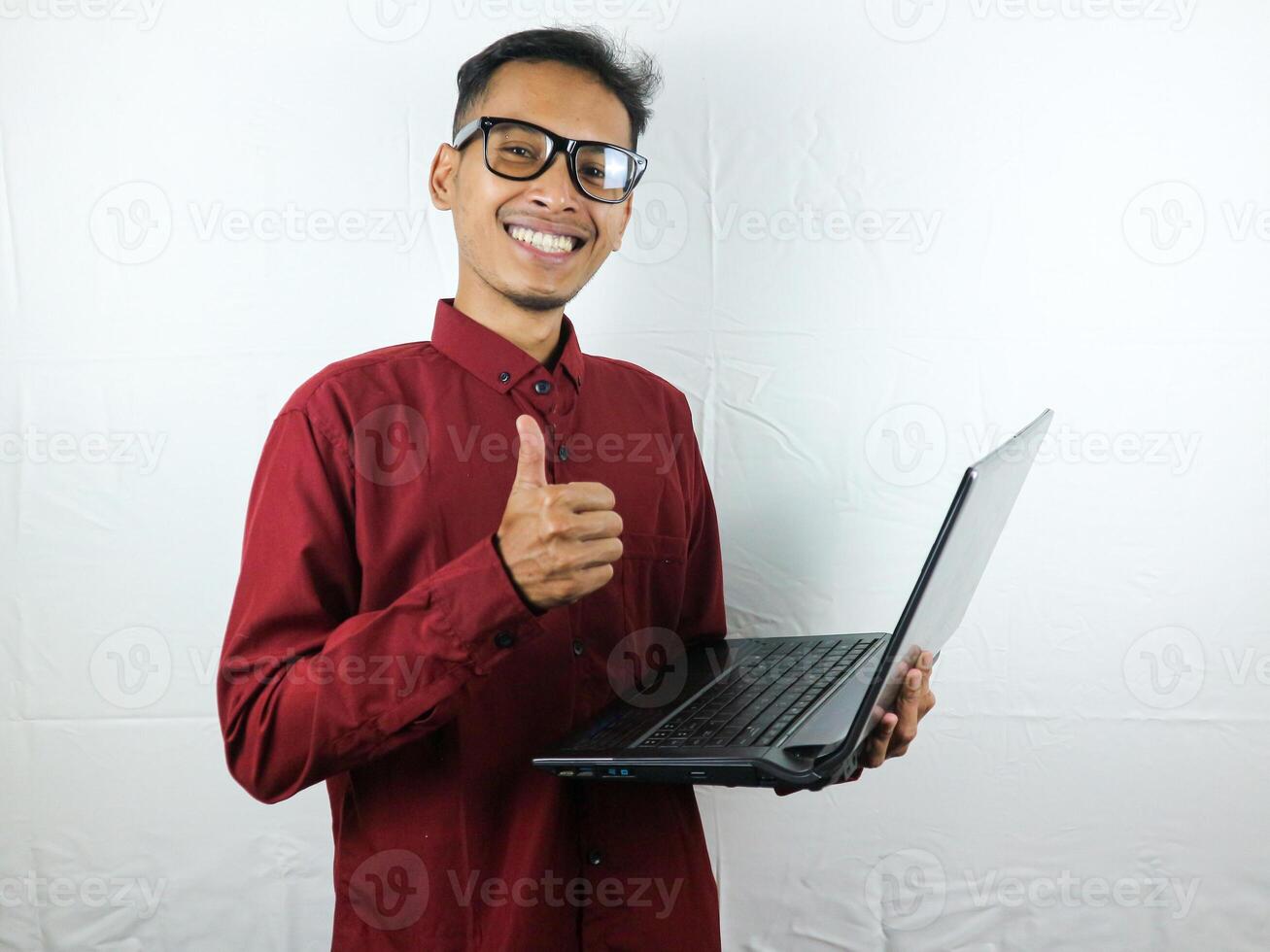 retrato de un asiático hombre vistiendo un rojo ropa participación un ordenador portátil con un sonriente cara expresión. foto