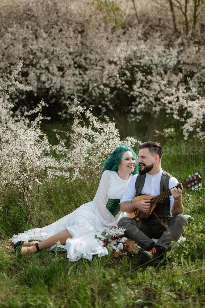 a bearded groom plays a stringed instrument and a girl sits in a spring meadow photo