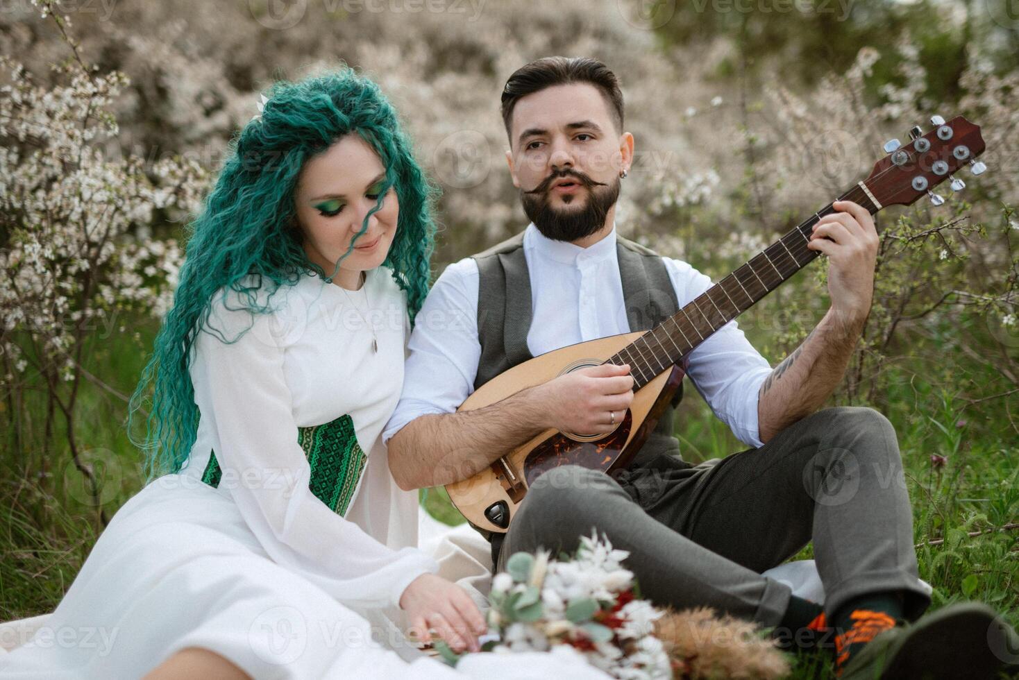 a bearded groom plays a stringed instrument and a girl sits in a spring meadow photo