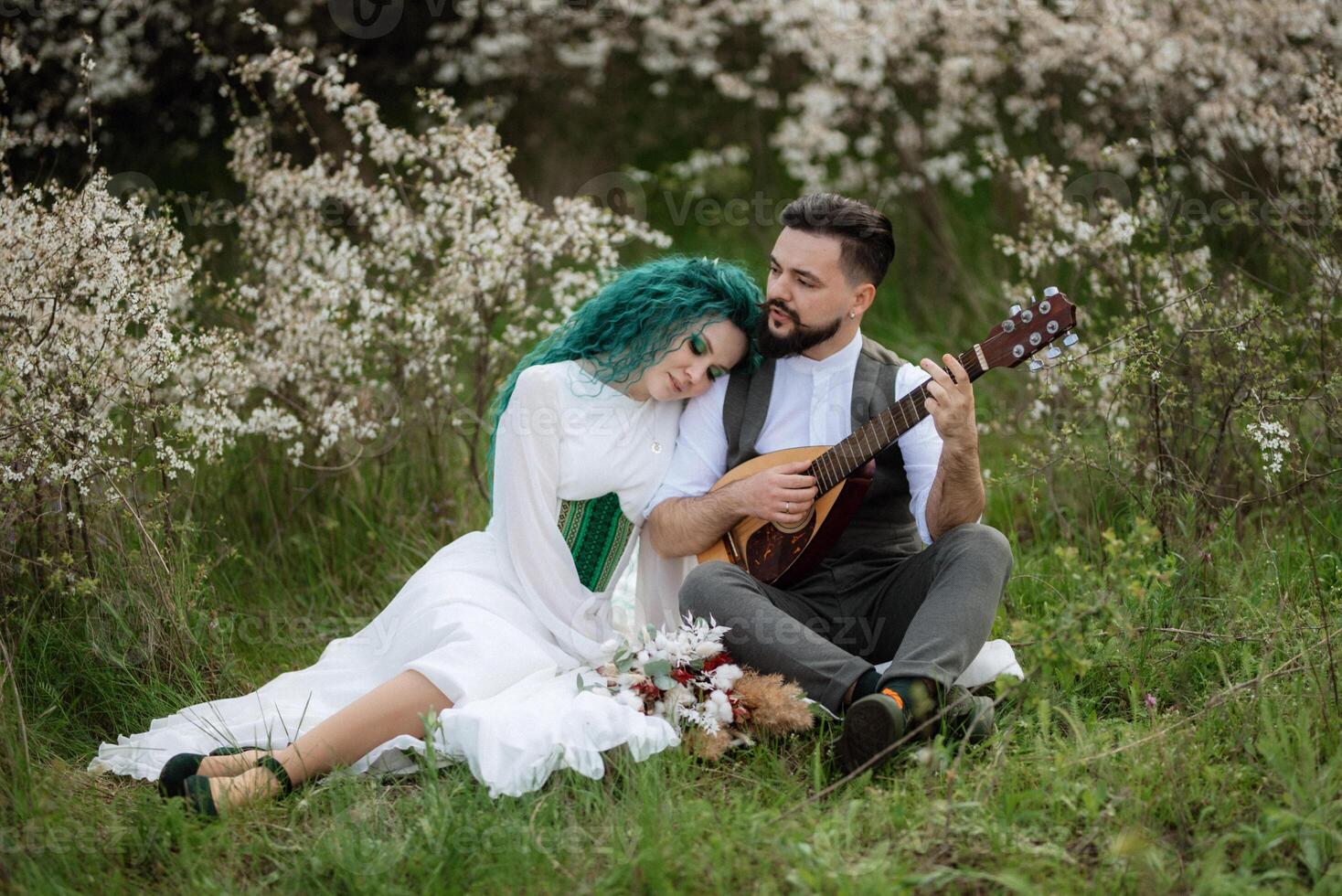 a bearded groom plays a stringed instrument and a girl sits in a spring meadow photo