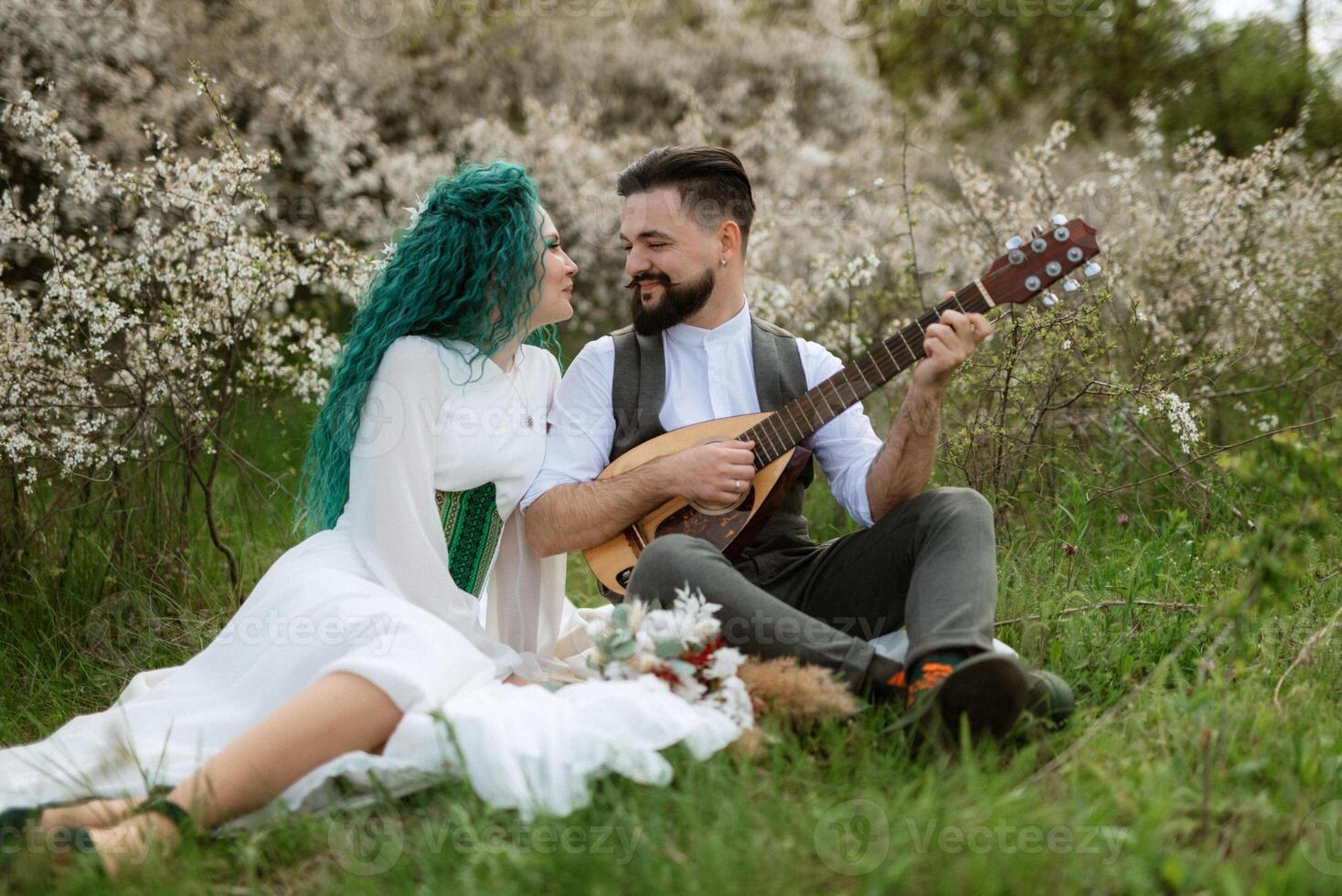 a bearded groom plays a stringed instrument and a girl sits in a spring meadow photo