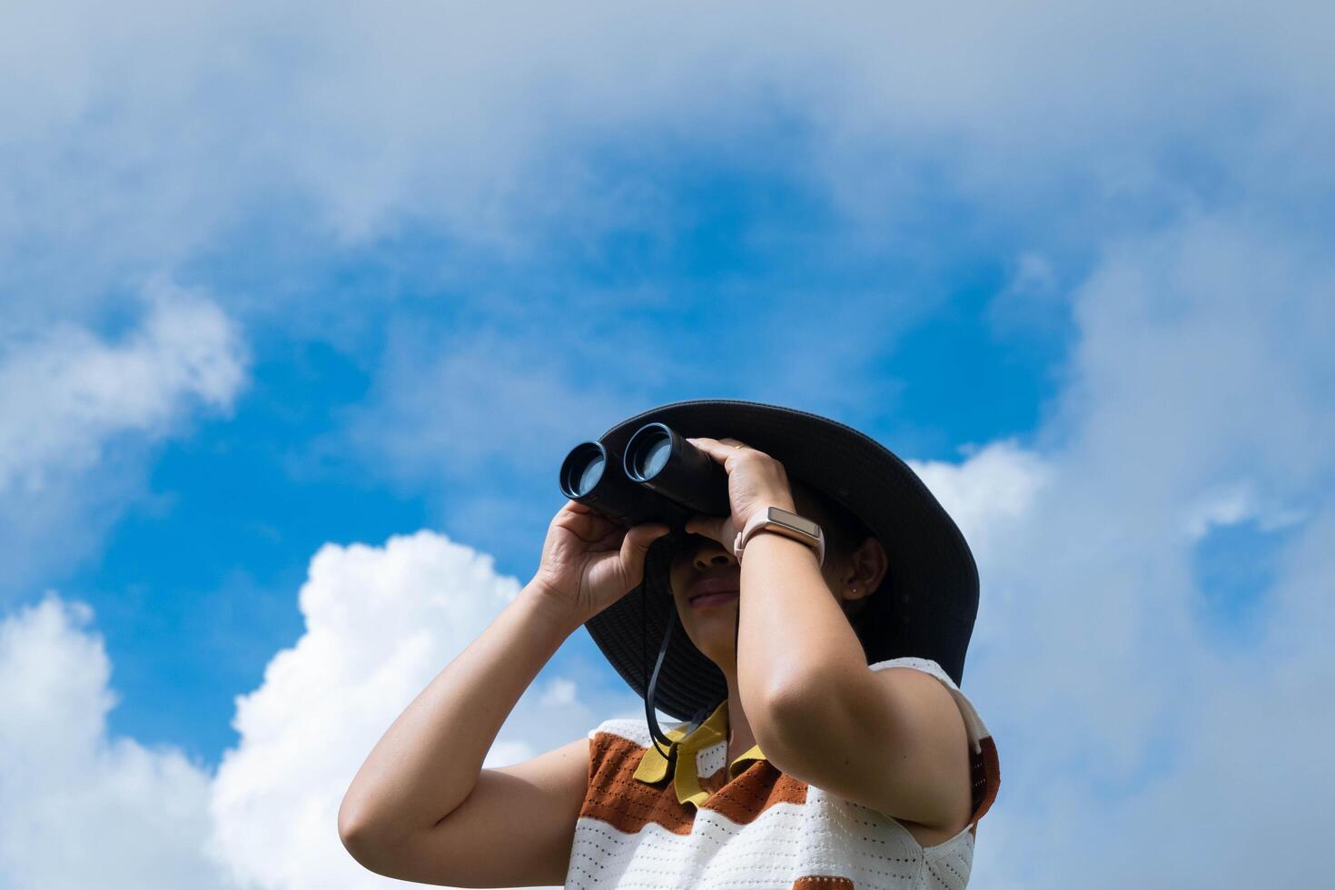 Young woman with binoculars on the mountain on a sunny day. Woman using binoculars when going hiking. Hiking woman uses binoculars to travel and has a happy smile. photo