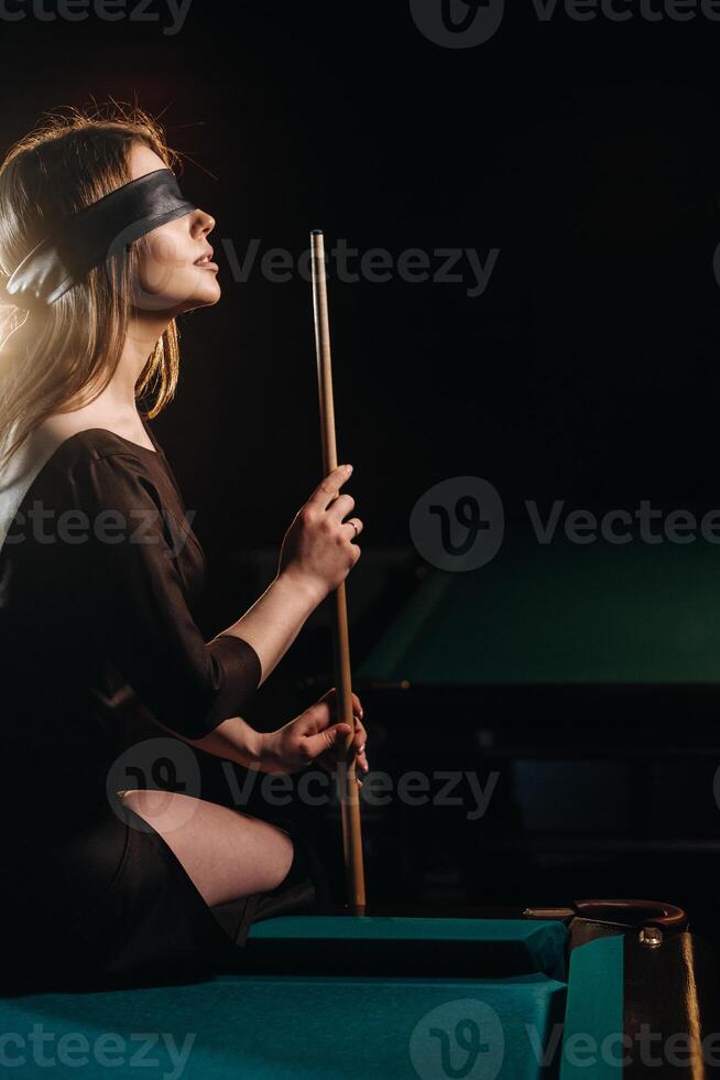 A girl with a blindfold and a cue in her hands is sitting on a table in a billiard club.Russian billiards photo