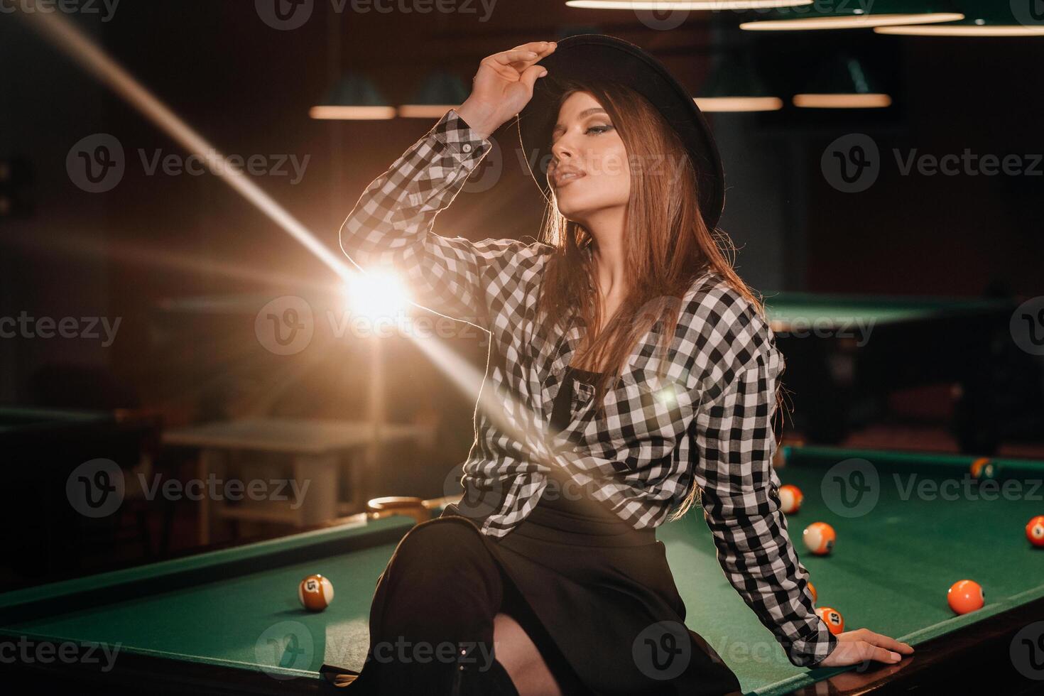 A girl in a hat in a billiard club sits on a billiard table.Playing pool photo