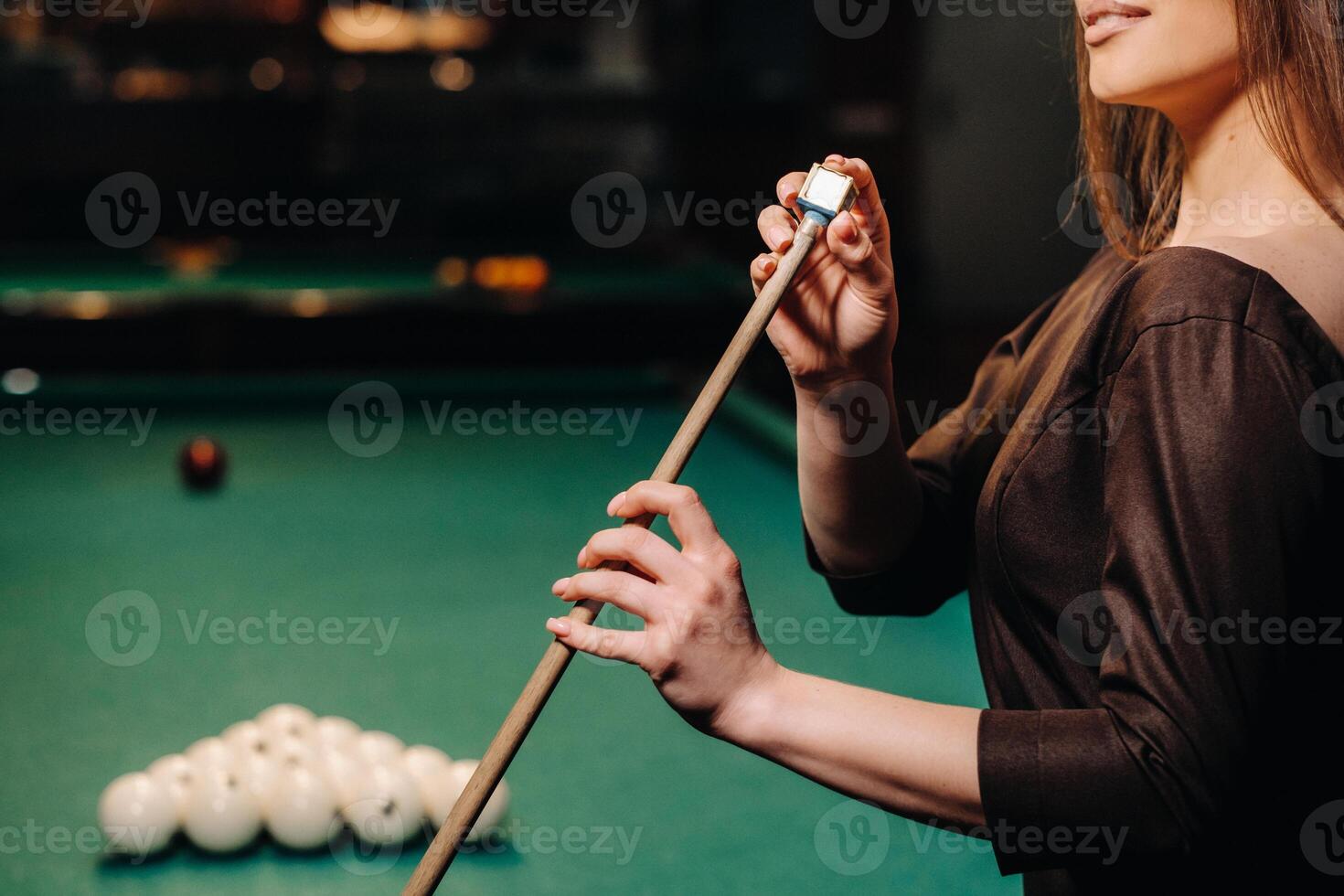 A girl in a dress stands with a cue in her hands and cleans it with chalk in a billiard club with balls in her hands.Playing billiards photo