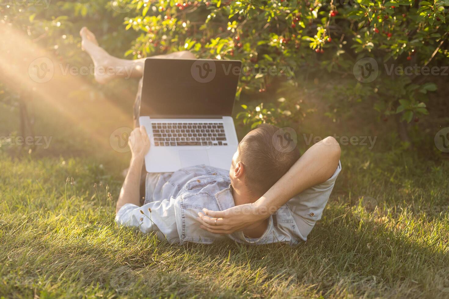 persona de libre dedicación hombre con ordenador portátil trabajando en jardín sentar en silla en césped al aire libre. joven blogger masculino trabajo en computadora en público parque procesos vídeo para social medios de comunicación contenido. sitio de trabajar, distancia trabajo. foto