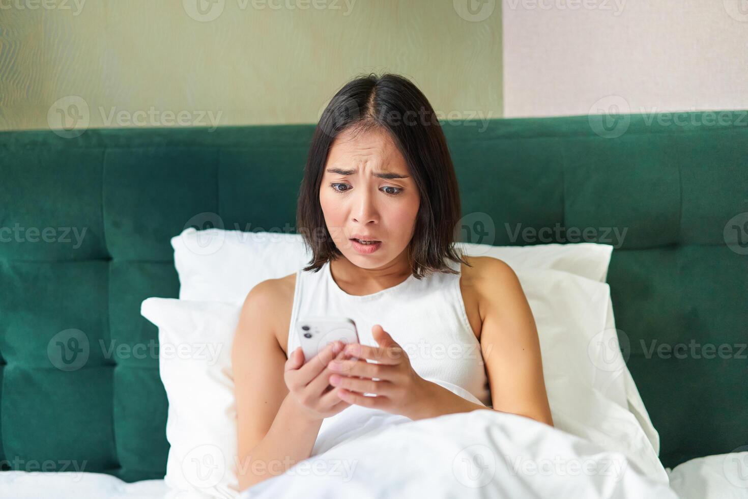Bedroom picture of asian woman lying in bed, looking scared and shocked at smartphone screen, reading message with concerned face photo