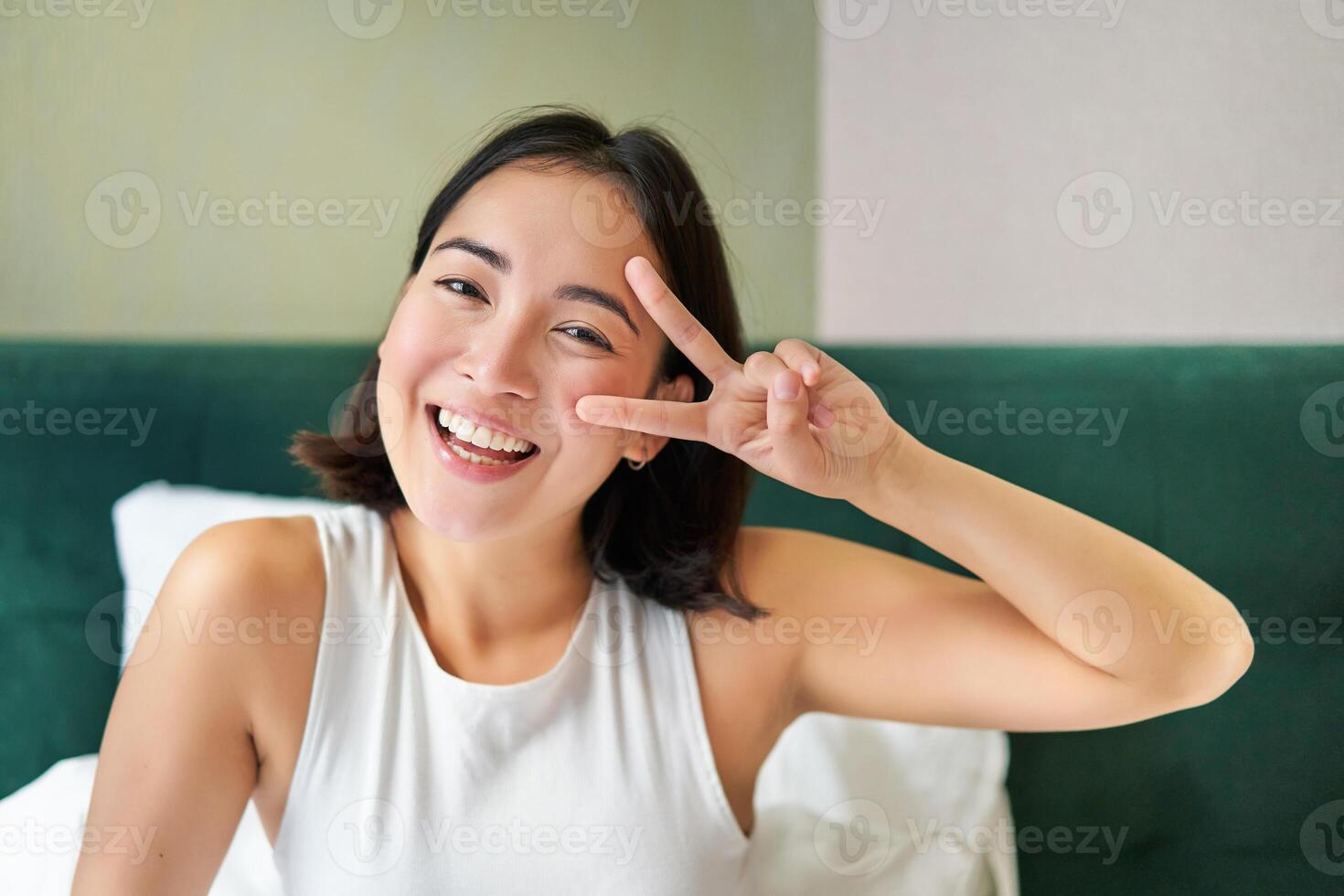 Positive asian woman lying in bed, showing peace sign, enjoys happy morning, waking up upbeat, staying in her bedroom photo