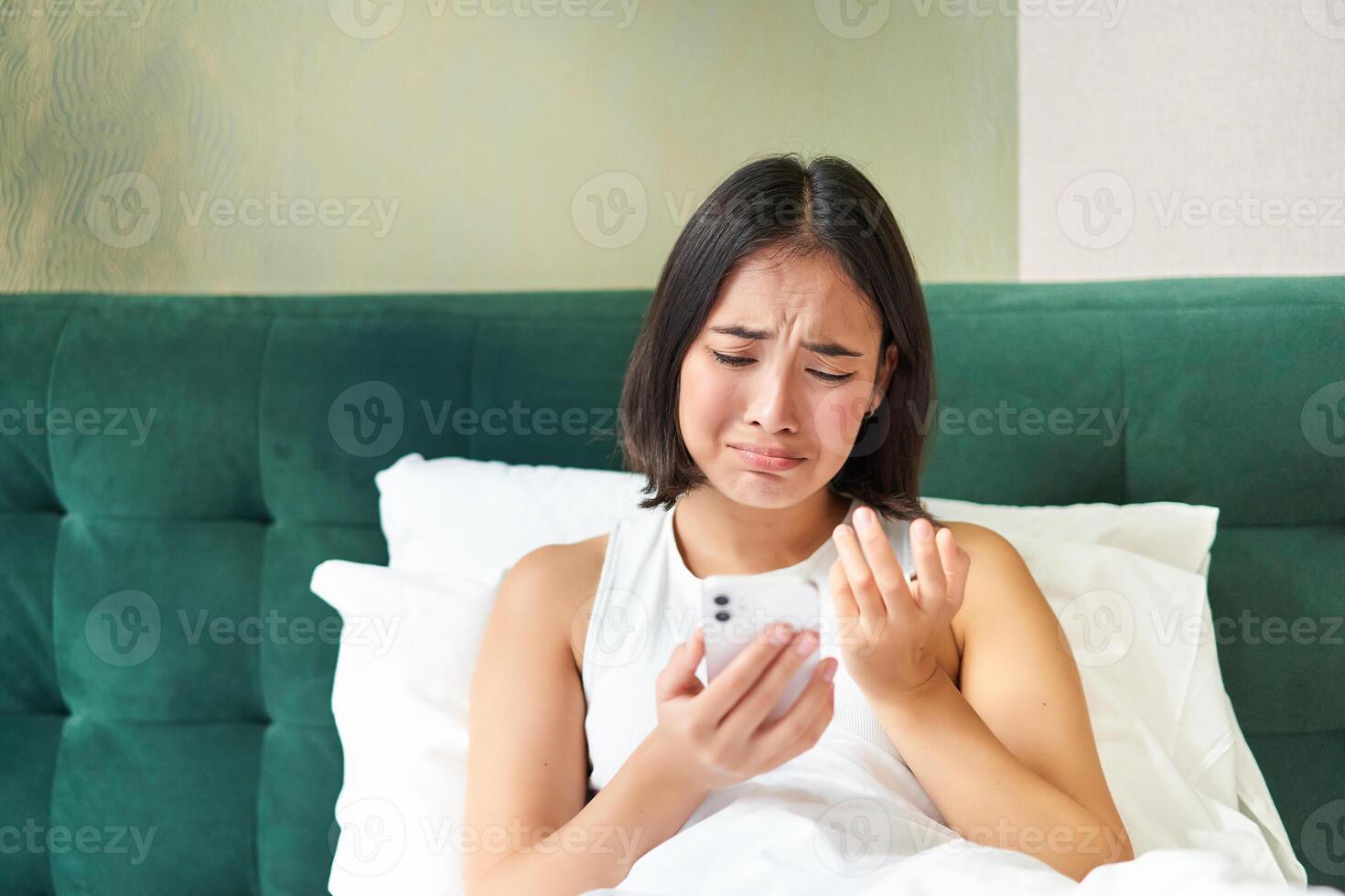 Portrait of sad frowning korean girl, lying in bed and looking at smartphone with upset, disappointed face expression, using mobile phone, complaining photo