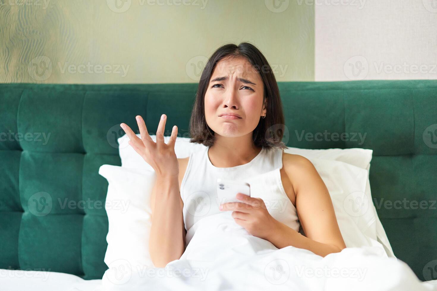 Disappointed asian girl, crying and whining, feeling upset, lying in bed on pillow, holding mobile phone, read upsetting bad news, waking up in awful mood photo