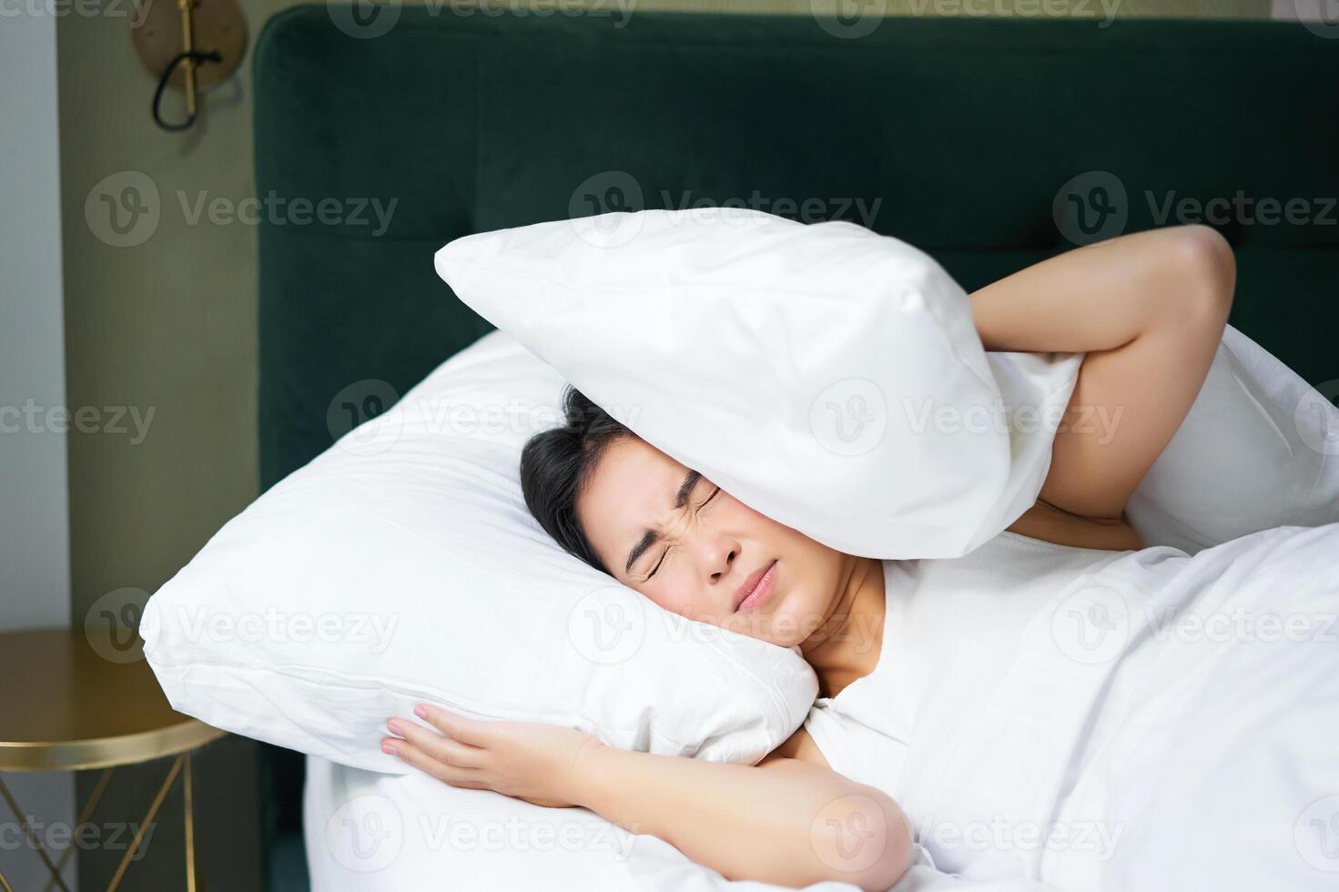 Insomnia. Asian girl shuts her ears with pillow as partner snoring. Woman lying in bed, bothered by noise at night, cannot sleep photo