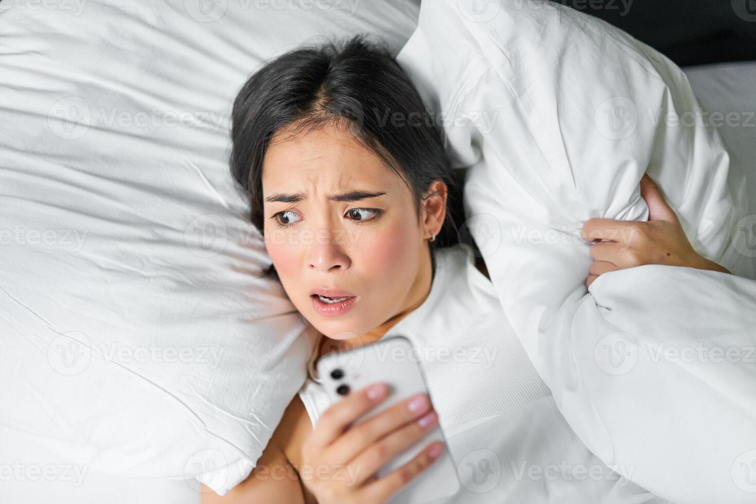 cerca arriba retrato de asiático niña acostado en cama, mirando a teléfono inteligente preocupado, despertar arriba tarde y curioso a su alarma reloj en móvil teléfono foto