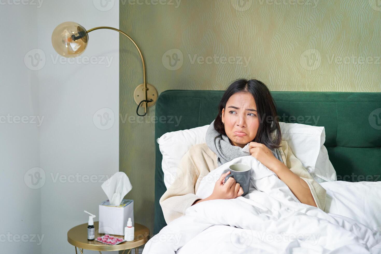 Covid-19, flu and vaccination concept. Korean girl lying in bed with cold, catching influenza, drinking hot tea and using prescribed medication photo