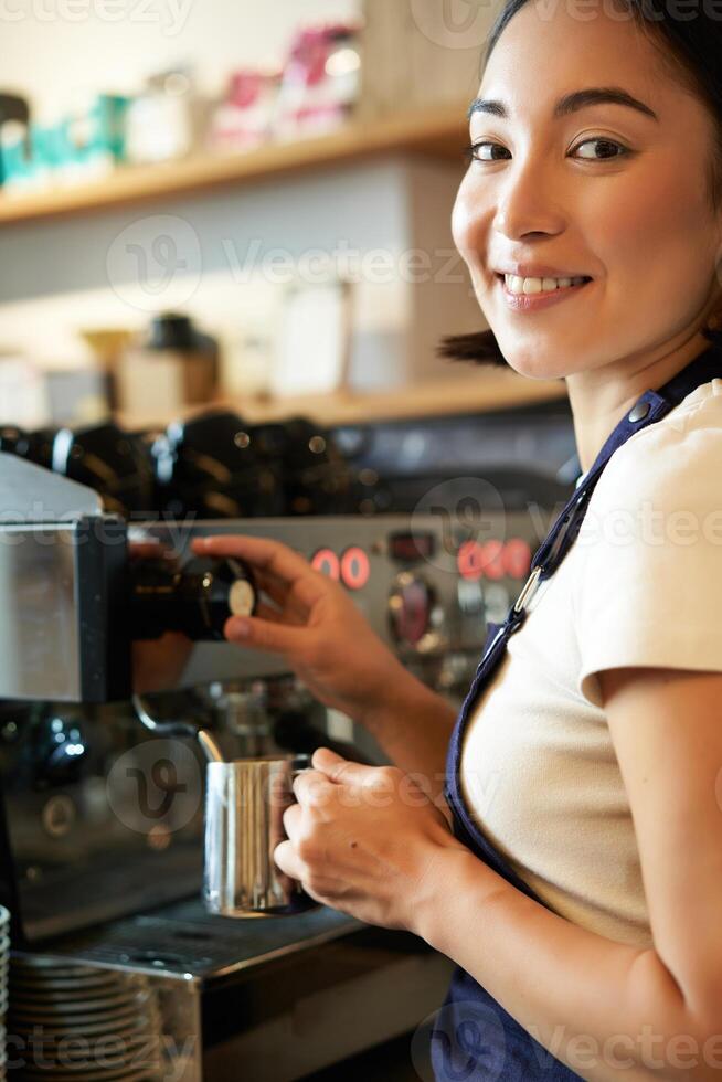 Vertical shot of barista, asian girl steaming milk for cappuccino, prepare latte for client, wearing blue apron, smiling happily, working in cafe photo