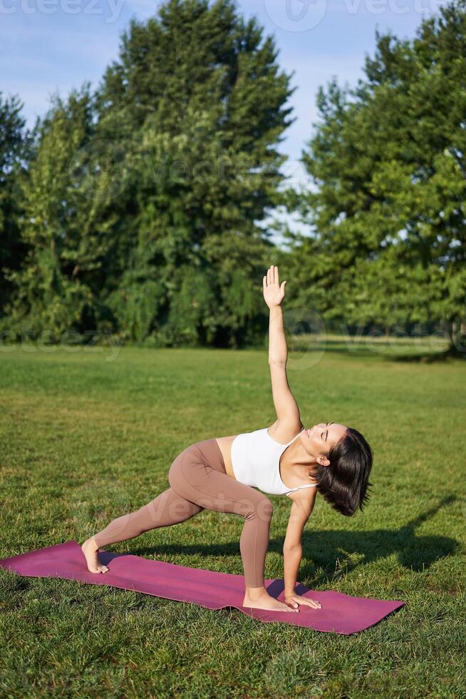 Vertical shot of young korean woman doing yoga training on rubber mat, making asana exercises on fresh air in park photo