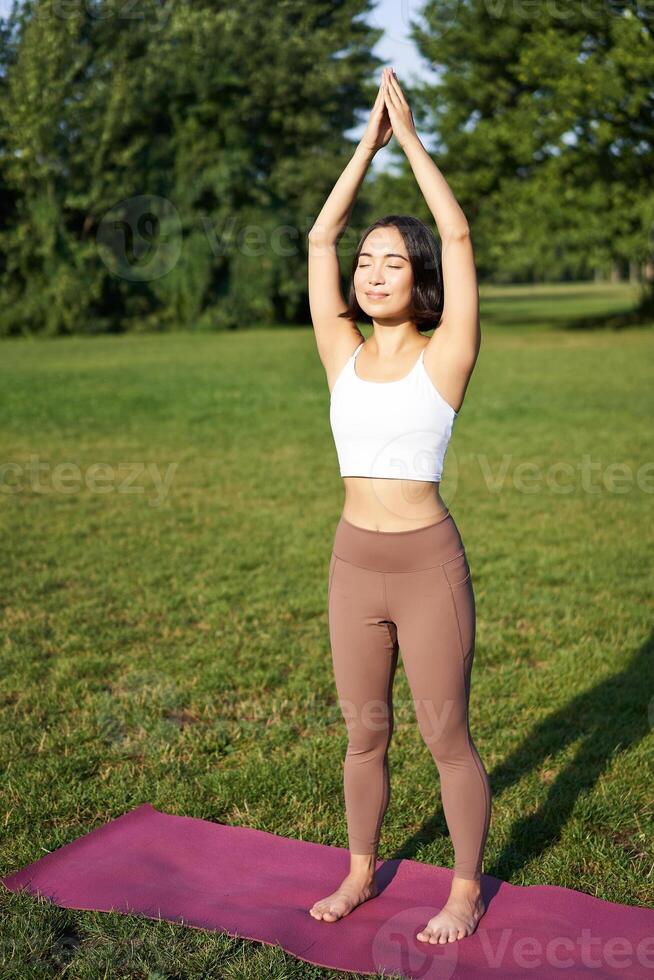 Smiling young fitness girl on rubber mat, workout in park, raising hands up in tree pose, doing yoga training on fresh green lawn photo