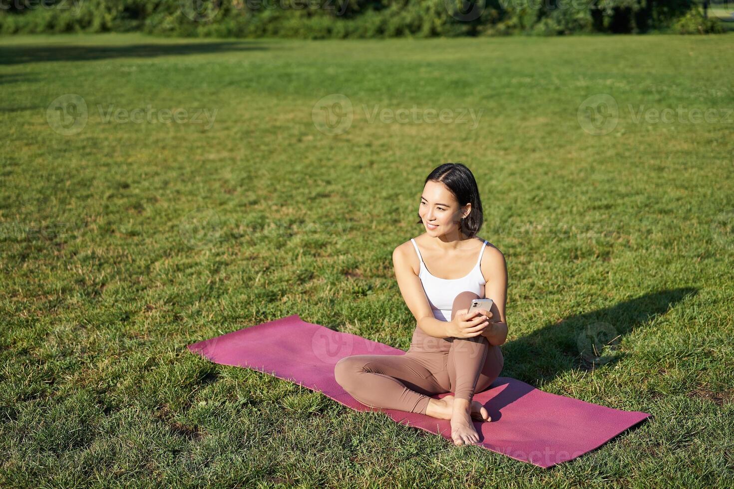 Woman in park, watching yoga video on smartphone, meditating on fresh air, sitting on rubber mat photo