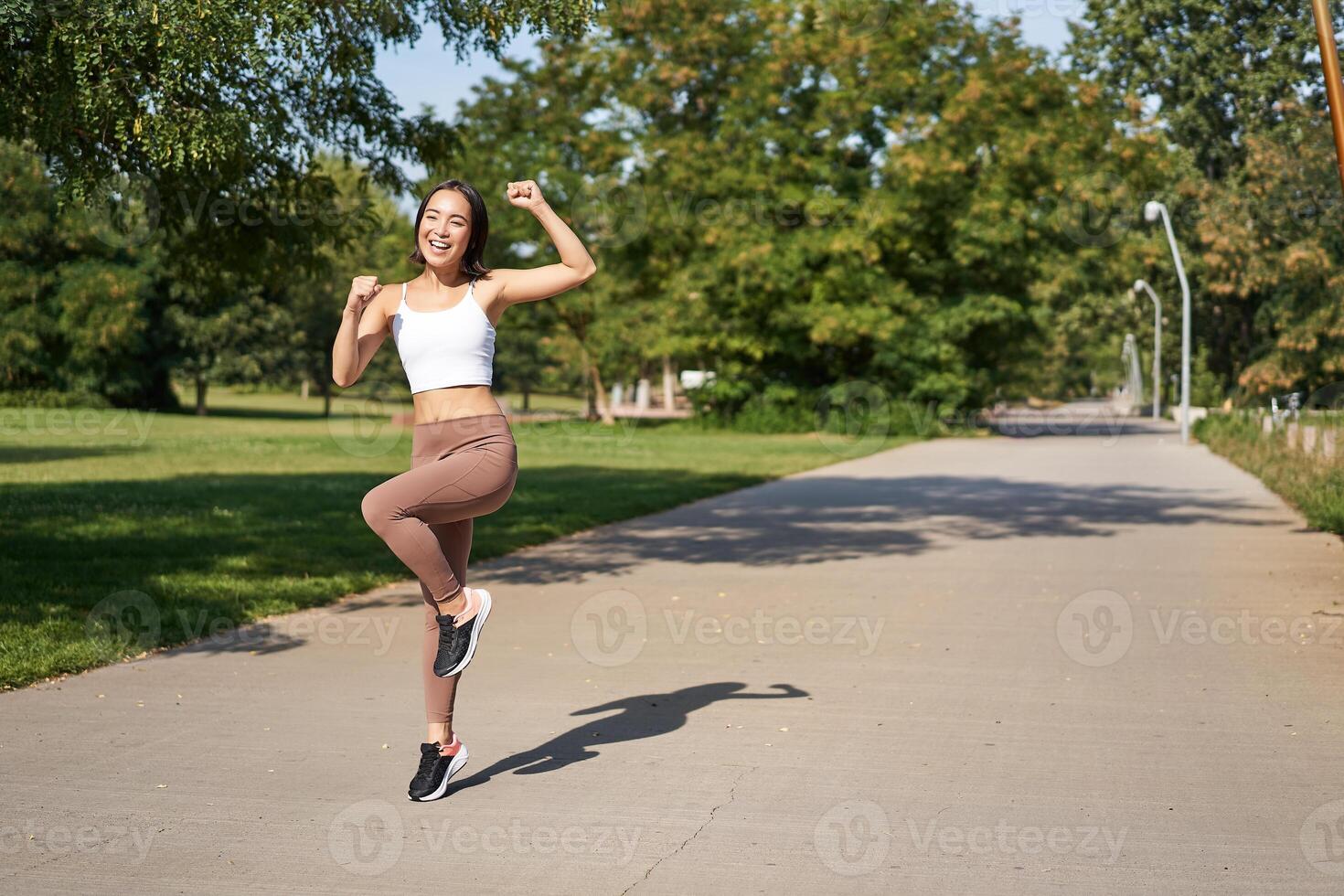 Excited young asian woman winning, finish running in park, saying yes, lifting hand up in triumph, celebrating victory or success photo