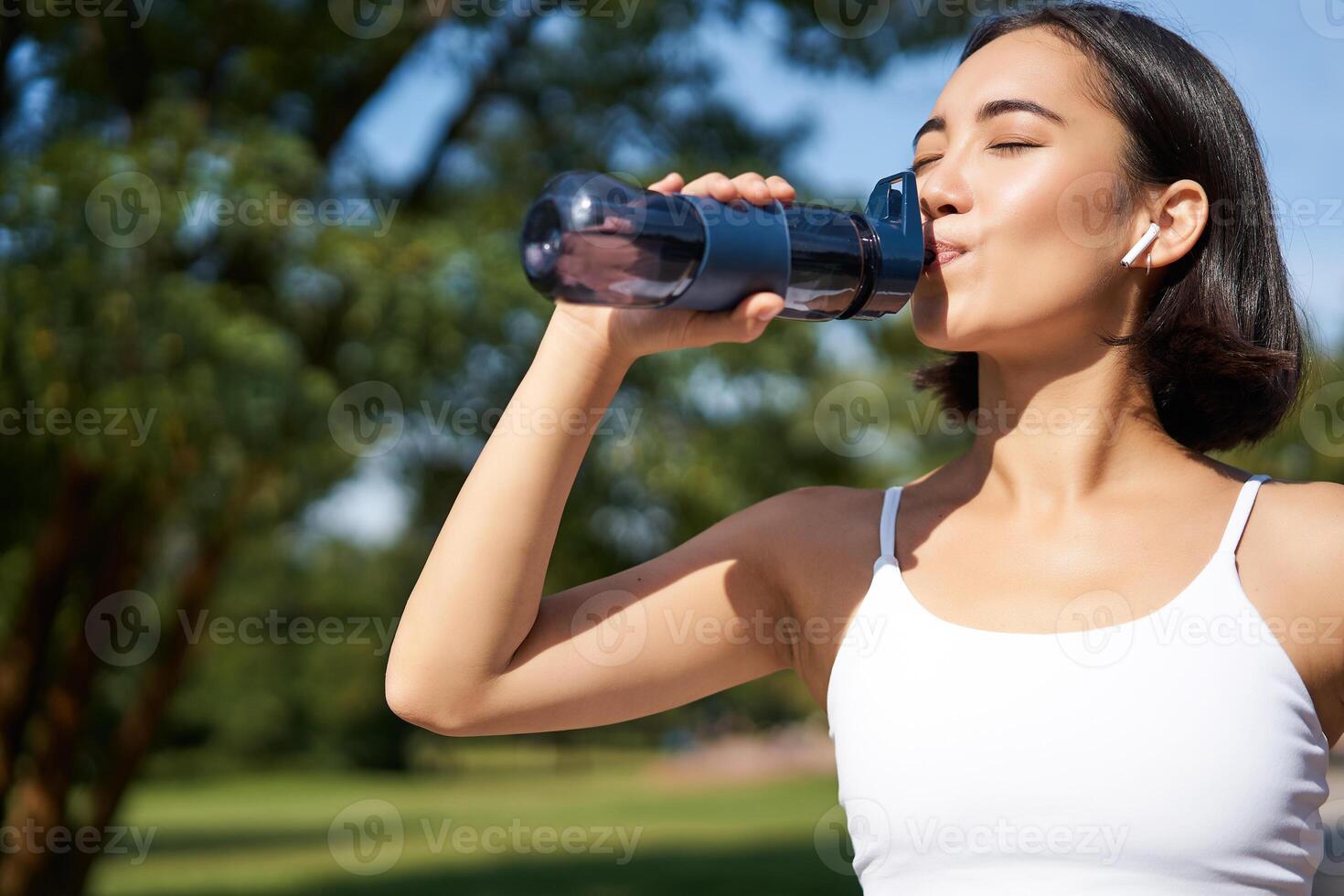 Fit asian sportswoman drinks water while running marathon. Fitness girl jogging in park, staying hydrated. photo