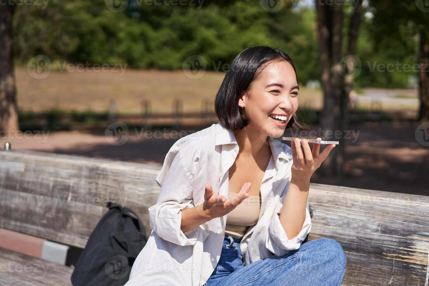 Portrait of happy asian girl records voice message, talking via speakerphone on smartphone, sitting relaxed on bench in park on sunny day photo