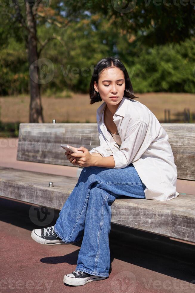 Portrait of asian girl sitting with smartphone feeling sad, looking gloomy and frustrated, waiting for a call outdoors in park photo