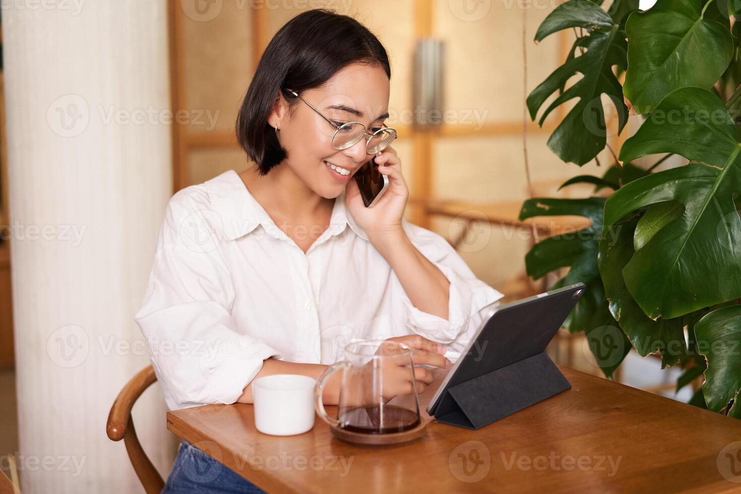 Woman in glasses, answer phone call in a cafe, drinking coffee, looking at work on tablet, working on remote from restaurant photo