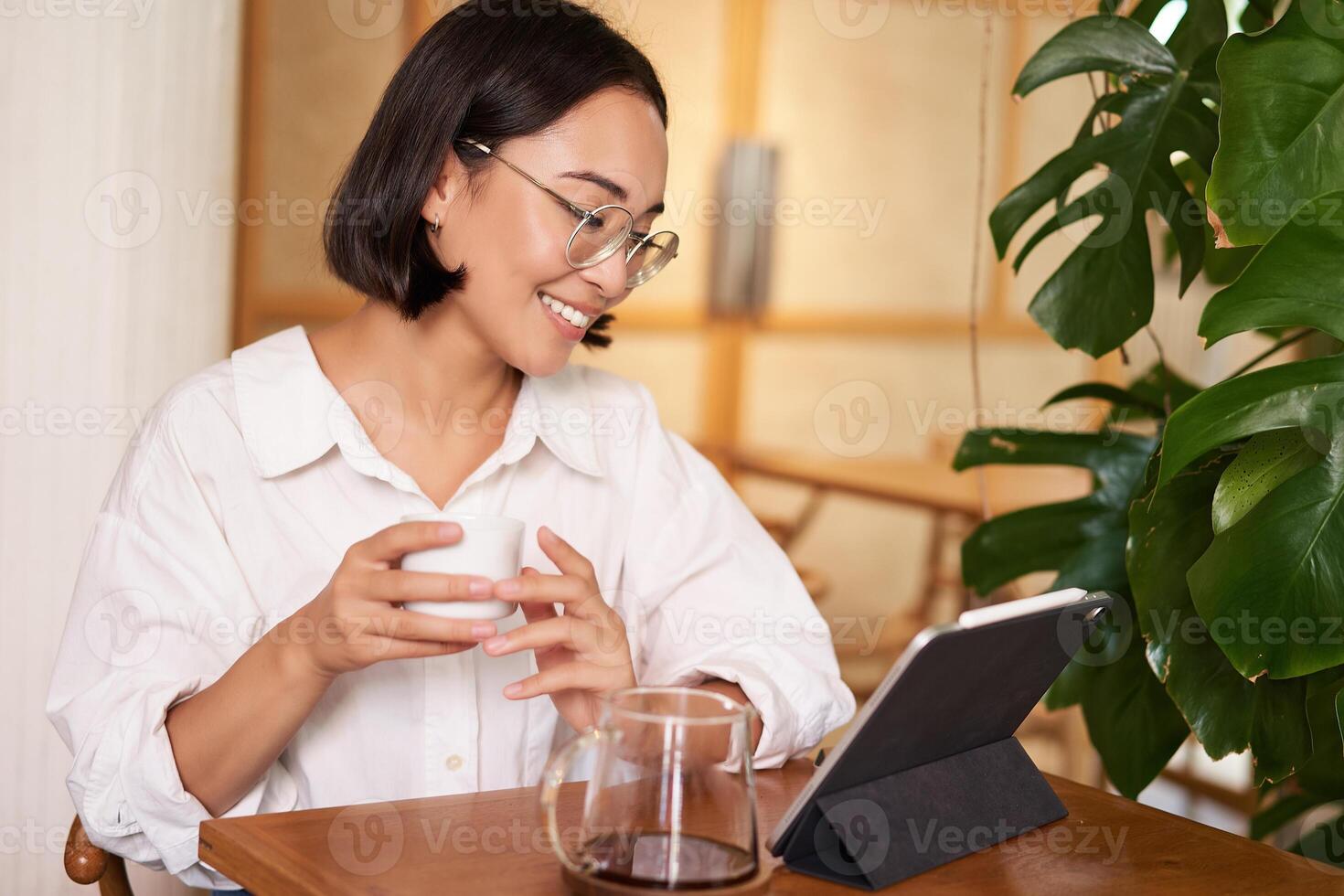 Asian woman in glasses, watching smth on digital tablet, drinking coffee in a cafe and smiling, working remotely photo