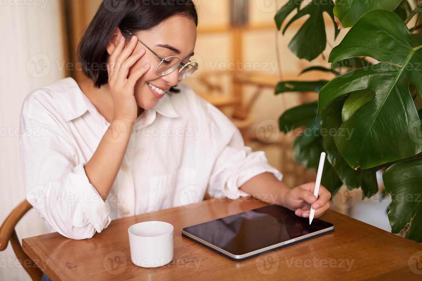 Young asian creative girl, sitting with digital tablet, drawing with graphic pen and smiling, doodling, drinking coffee in cafe photo