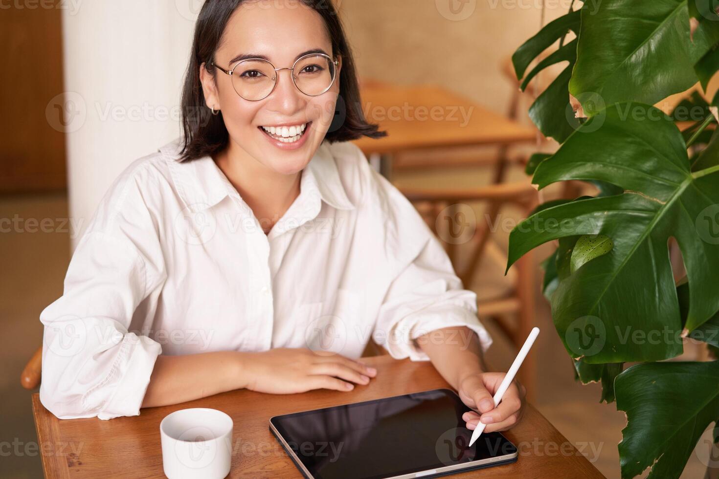 Smiling young female creator, creative girl drawing on digital tablet with graphic pen, drinking coffee in cafe photo
