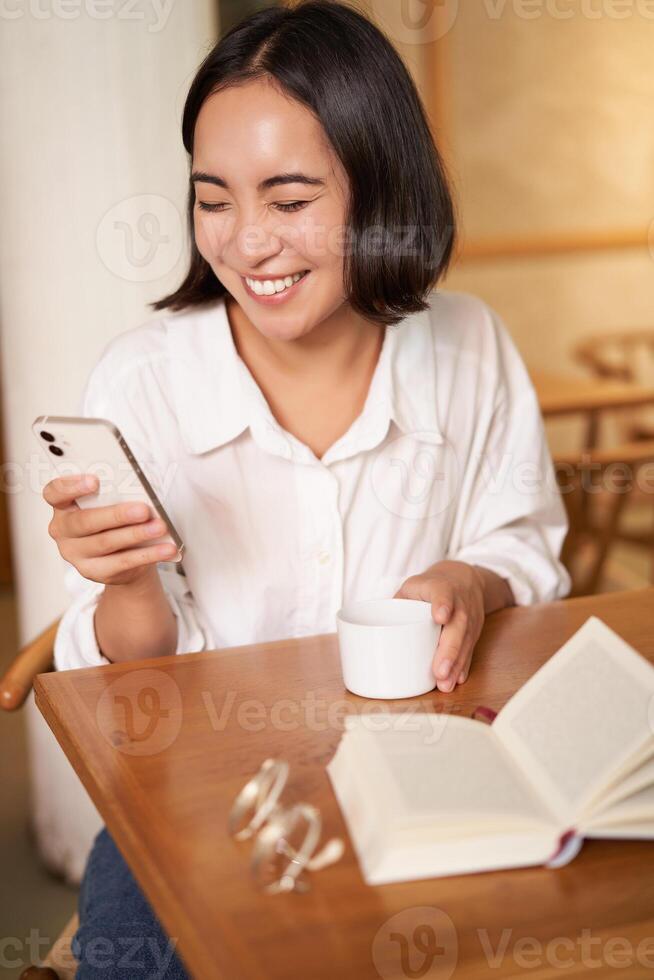 Happy asian woman sits in cafe with cup of coffee and book, answer video call on smartphone, laughing and talking via camera on mobile phone photo