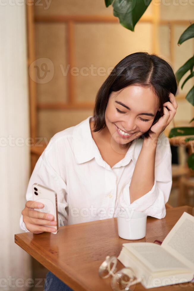 Happy asian woman sits in cafe with cup of coffee and book, answer video call on smartphone, laughing and talking via camera on mobile phone photo