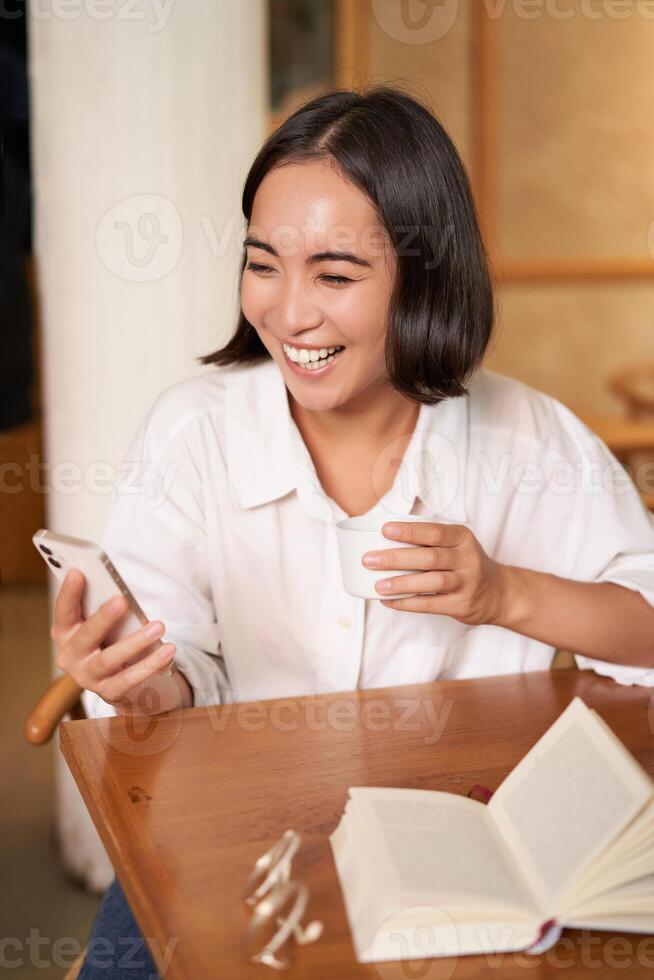 Happy asian woman sits in cafe with cup of coffee and book, answer video call on smartphone, laughing and talking via camera on mobile phone photo