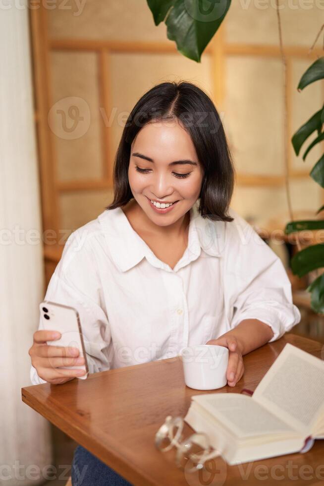 Happy asian woman sits in cafe with cup of coffee and book, answer video call on smartphone, laughing and talking via camera on mobile phone photo
