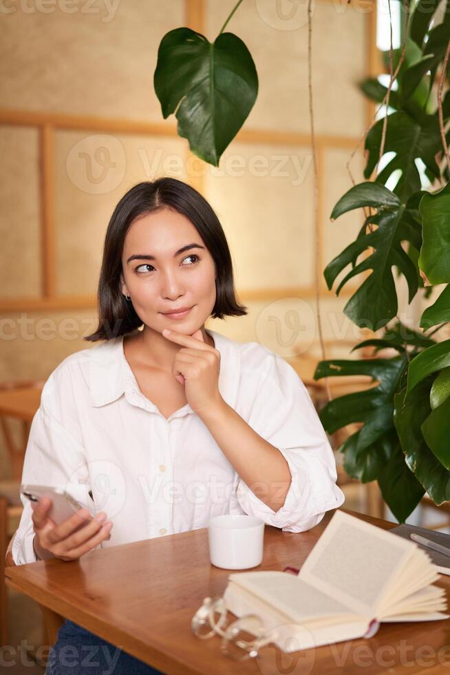 hermosa joven mujer, 25 años viejo, pensamiento, participación teléfono inteligente y mirando considerado, sentado en cafetería, decidiendo algo foto