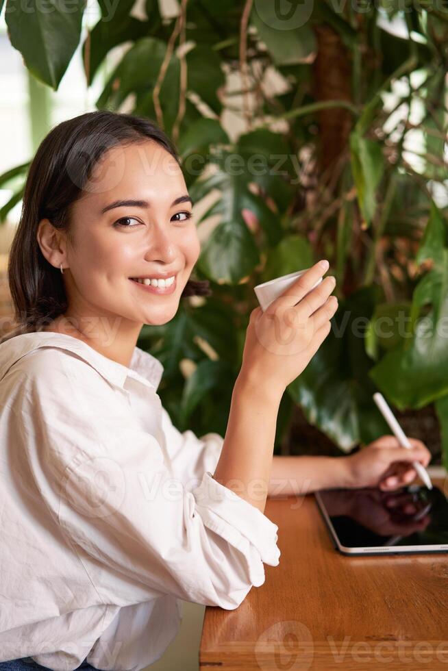 Vertical shot of young asian woman with digital tablet and graphic pen, drawing, scratching while sitting in cafe with cup of coffee photo