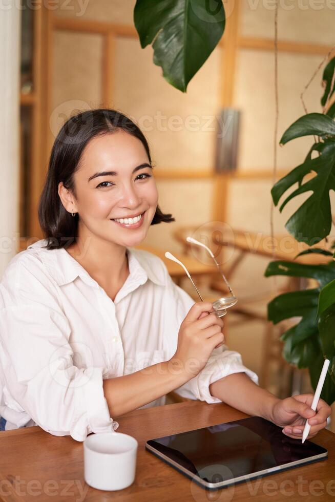 Beautiful asian girl sitting in cafe with cup of coffee, drawing on digital tablet with graphic pen, doddling and making scatches photo