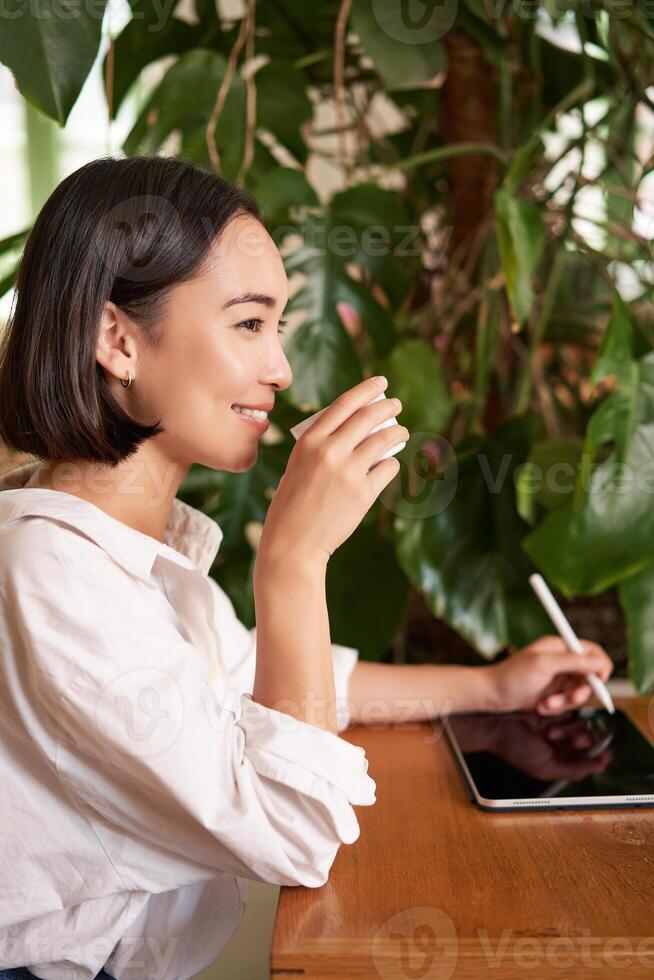 Beautiful asian girl sitting in cafe with cup of coffee, drawing on digital tablet with graphic pen, doddling and making scatches photo