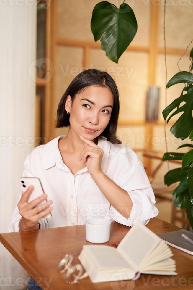 hermosa joven mujer, 25 años viejo, pensamiento, participación teléfono inteligente y mirando considerado, sentado en cafetería, decidiendo algo foto