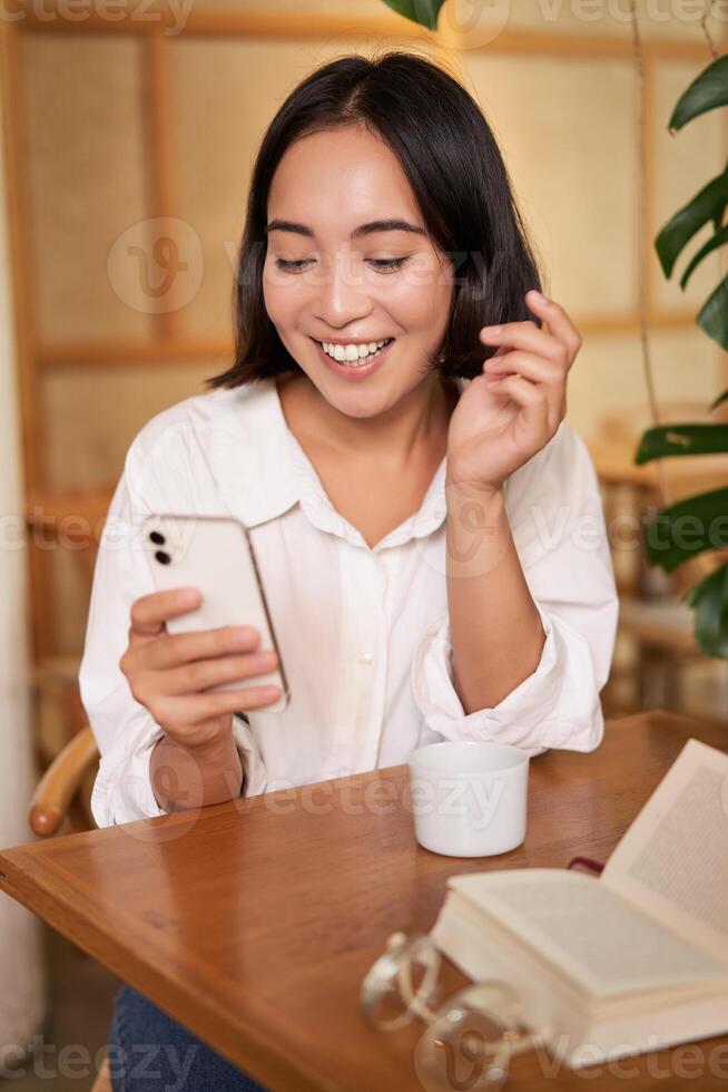Portrait of modern girl sits with coffee in cafe, smiling while looking at smartphone, reading book in restaurant and using mobile phone app photo