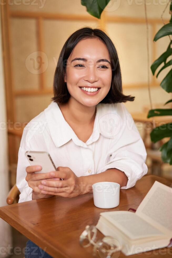 Portrait of modern girl sits with coffee in cafe, smiling while looking at smartphone, reading book in restaurant and using mobile phone app photo