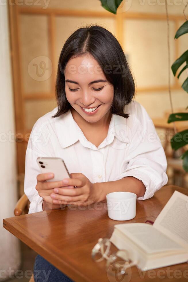 Portrait of modern girl sits with coffee in cafe, smiling while looking at smartphone, reading book in restaurant and using mobile phone app photo