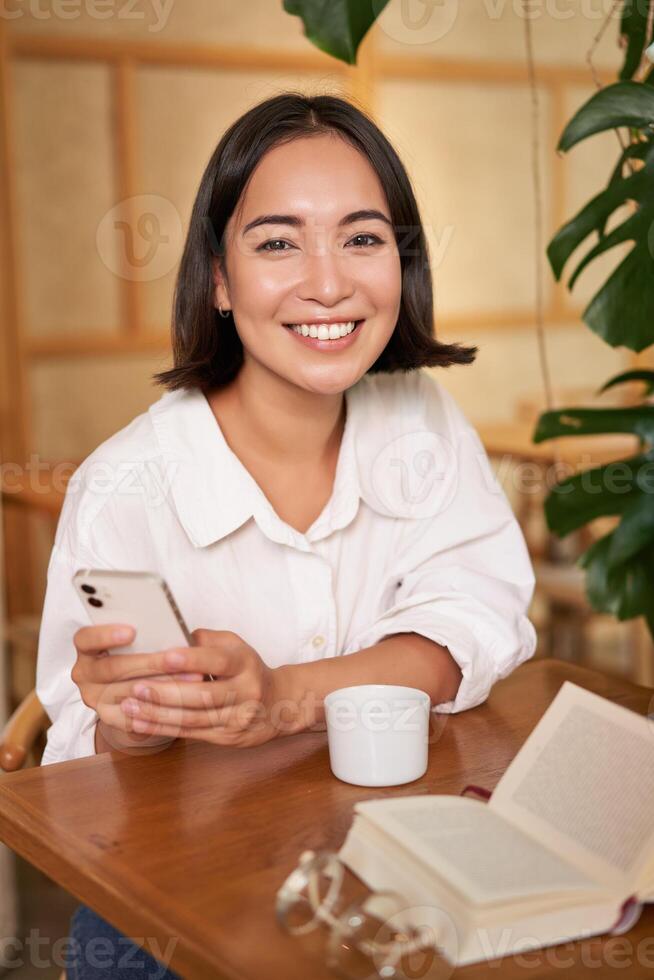 Portrait of korean girl in cafe, drinking coffee, using mobile phone and reading a book in cozy place photo