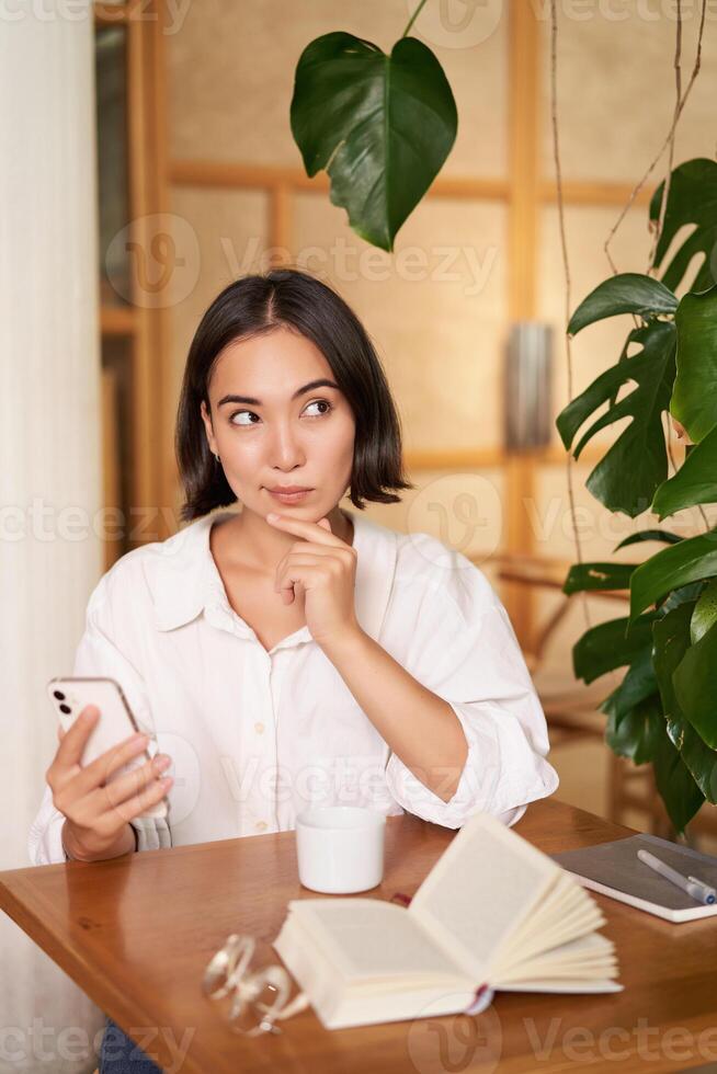 hermosa joven mujer, 25 años viejo, pensamiento, participación teléfono inteligente y mirando considerado, sentado en cafetería, decidiendo algo foto