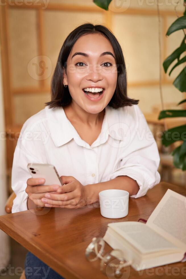 Portrait of korean girl in cafe, drinking coffee, using mobile phone and reading a book in cozy place photo