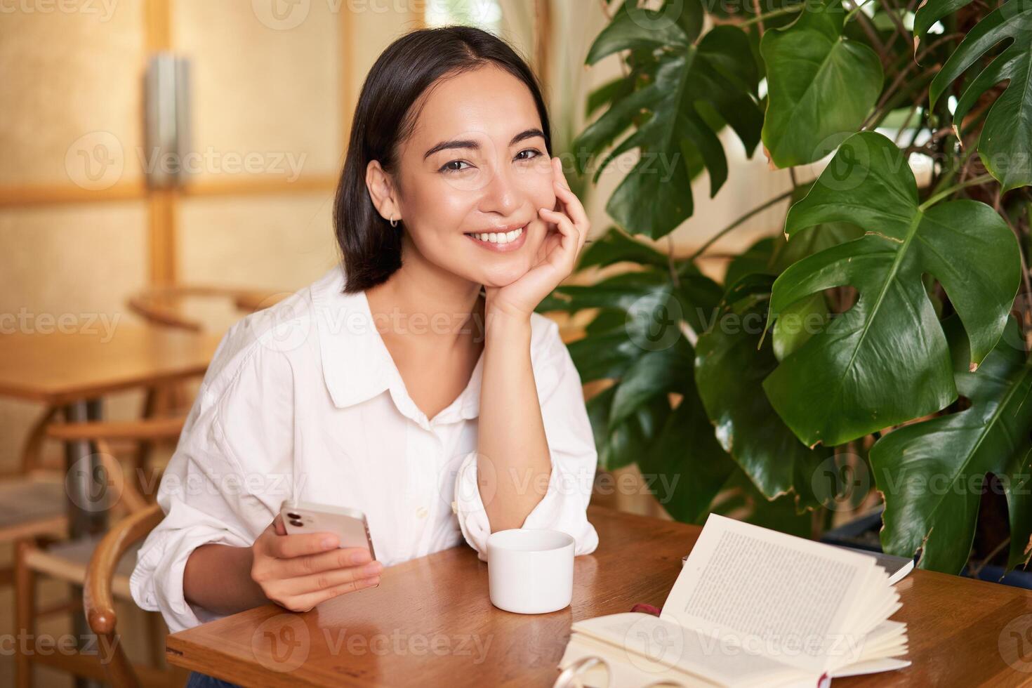 Beautiful young woman with smartphone, drinking coffee in cafe and smiling, relaxing with favorite book photo