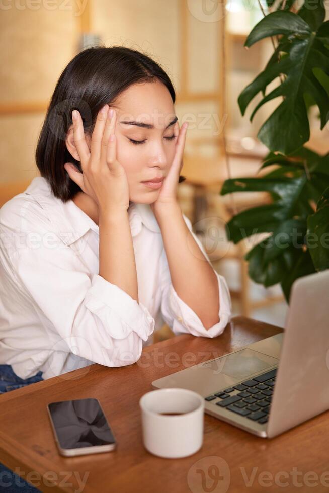 Vertical shot of asian girl looking tired at laptop screen, bored of working, sitting in cafe sleepy and exhausted photo